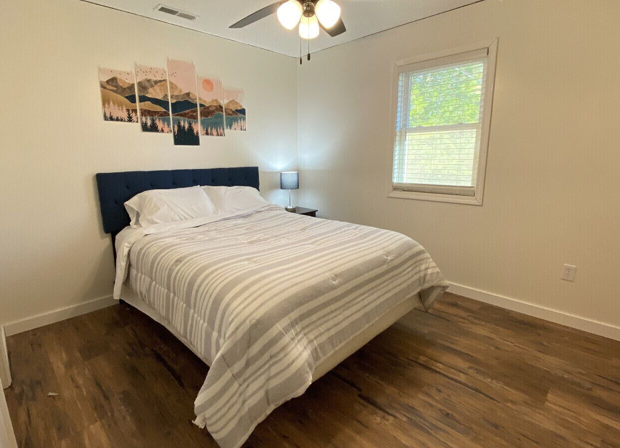 A bedroom with a bed, wooden flooring, a ceiling fan, and a window.