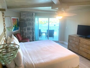 Ein Hotelzimmer mit einem großen Bett, einem Fernseher, einer Holzkommode, einem Sessel und einem Balkon mit Blick.