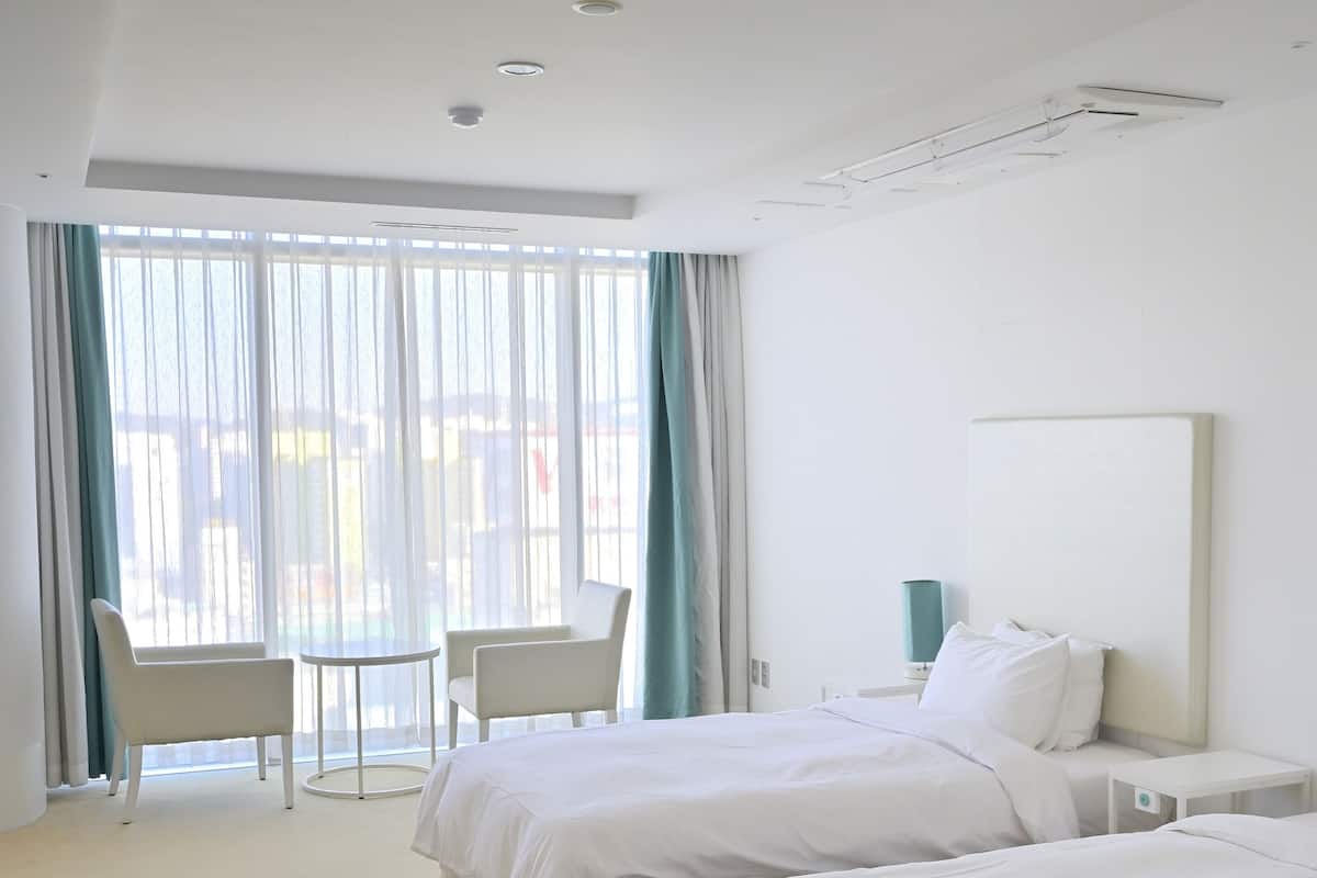 A hotel room with two beds, a chair, a small table, and a large window with curtains.