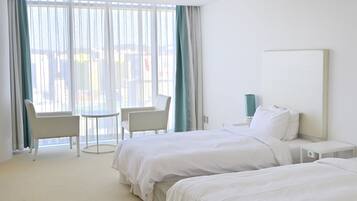 Habitación de hotel con dos camas, una silla, una mesita pequeña y un ventanal grande con cortinas.