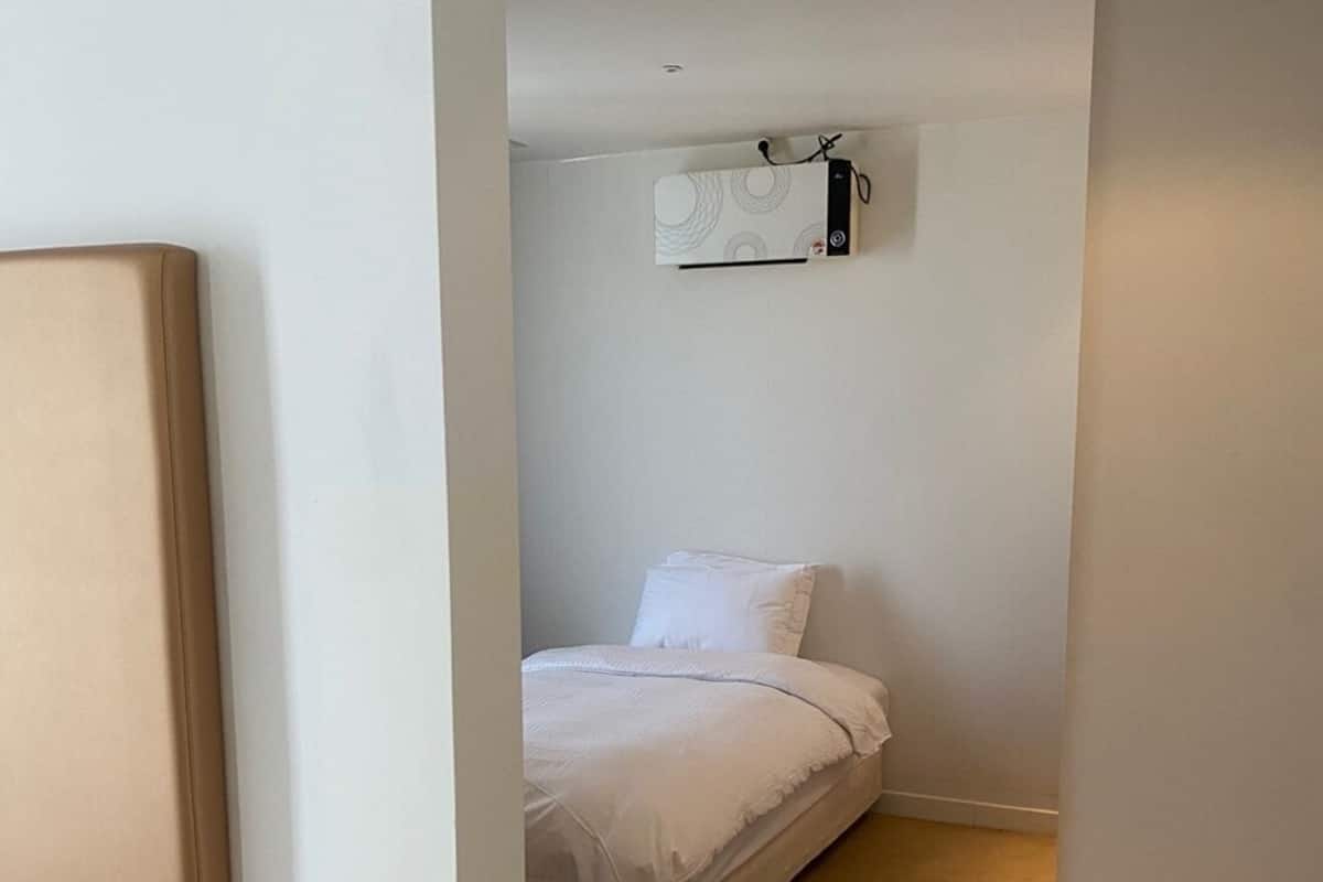 A narrow hallway with a bed at the end, a white wall, and a ceiling-mounted air conditioning unit.