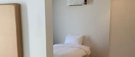 A narrow hallway with a bed at the end, a white wall, and a ceiling-mounted air conditioning unit.