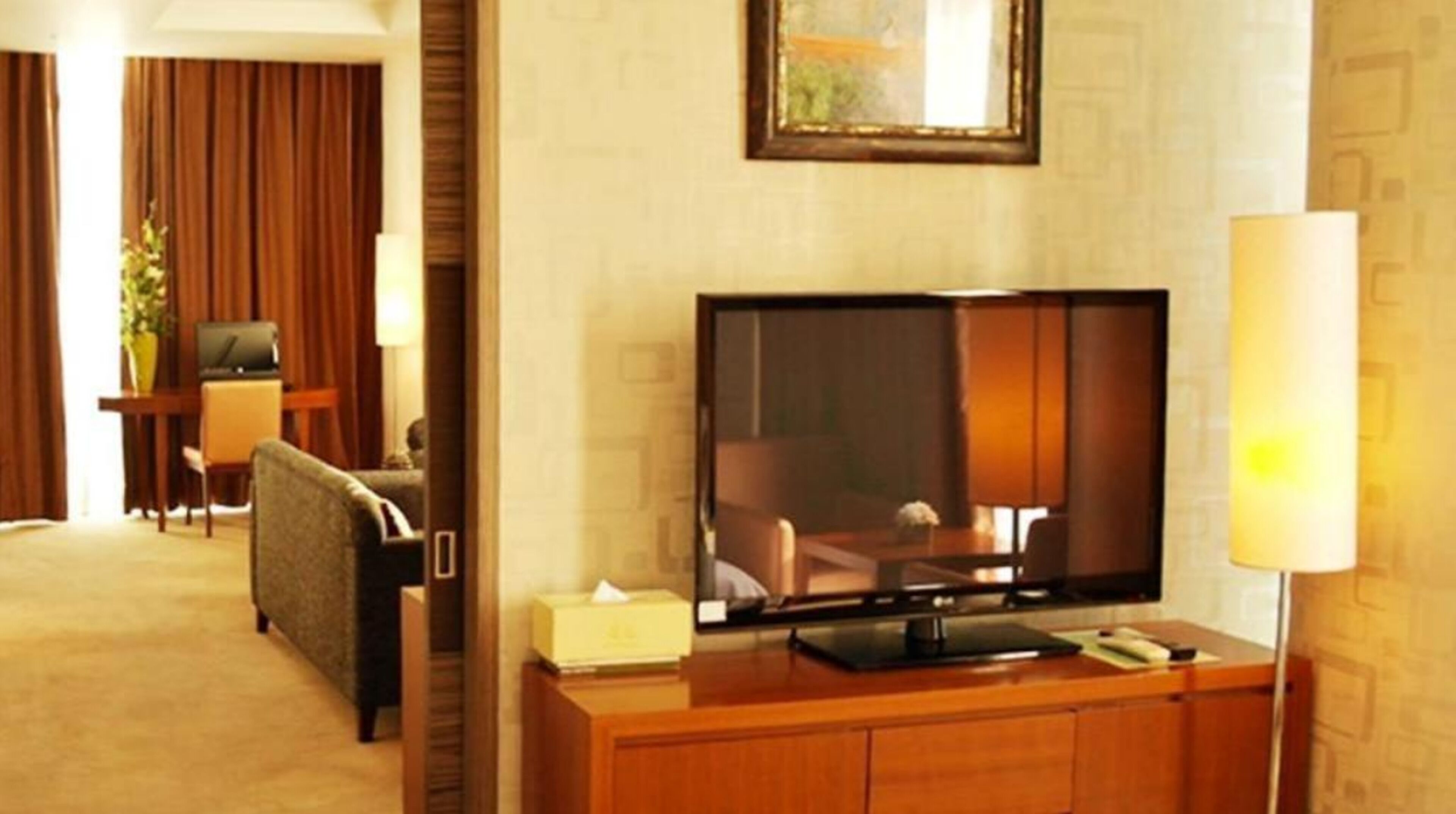 A hotel room with a flat-screen TV on a wooden console, a sofa, a desk with a lamp, and a large window with curtains.