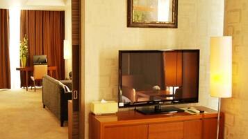 A hotel room with a flat-screen TV on a wooden console, a sofa, a desk with a lamp, and a large window with curtains.
