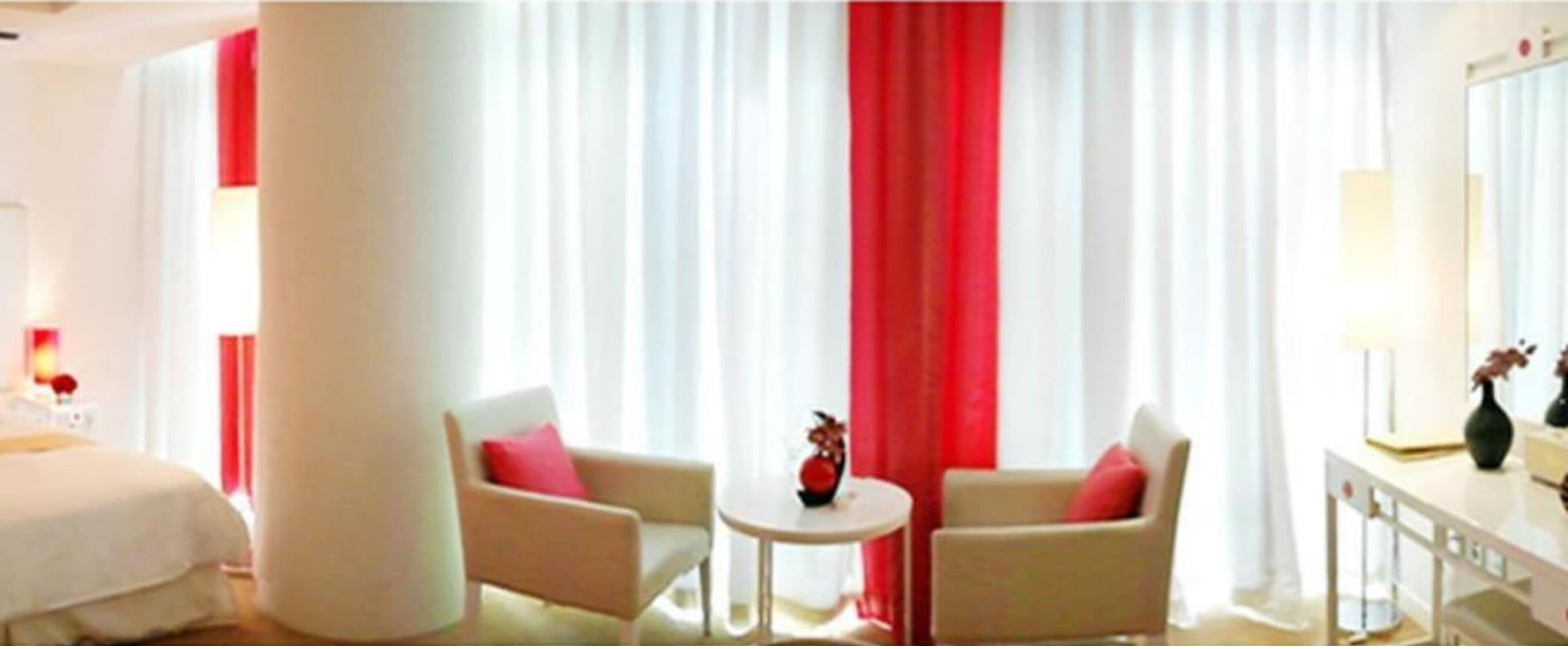Ein Hotelzimmer mit einem Bett, zwei Sesseln, einem kleinen runden Tisch und einem großen Fenster mit roten Vorhängen.
