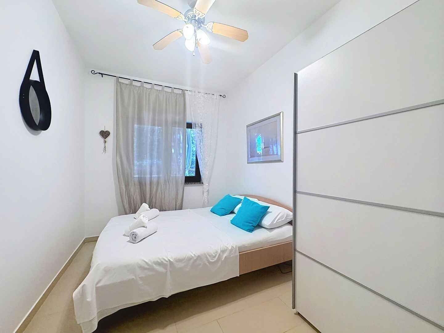 A bedroom with a bed, a ceiling fan, a window with curtains, a wall-mounted picture, and a wardrobe.