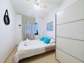 A bedroom with a bed, a ceiling fan, a window with curtains, a wall-mounted picture, and a wardrobe.