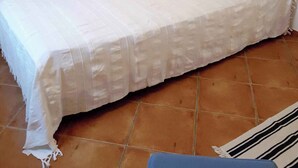 Un dormitorio con cama, ropa de cama blanca, cabecera, pared con cuadros, lámpara y ventana con persianas.