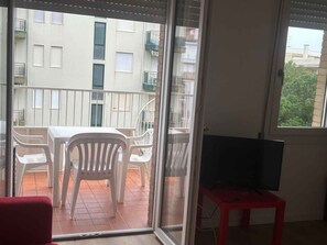 A balcony with a table and chairs, a red chair, a television on a stand, and a view of a building with balconies.