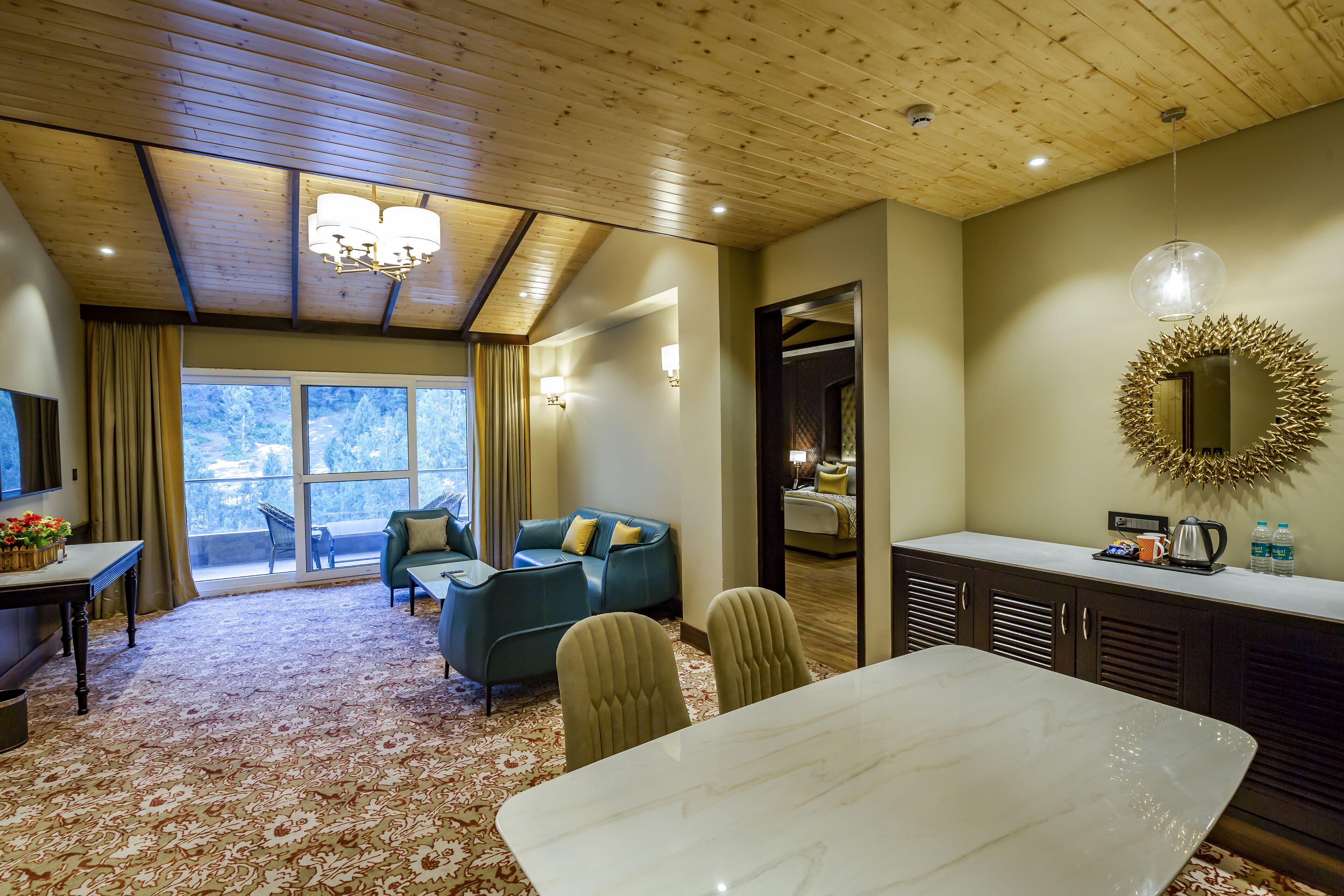 A hotel room with a wooden ceiling, a dining table with chairs, a sofa, a TV, and a large window with curtains.