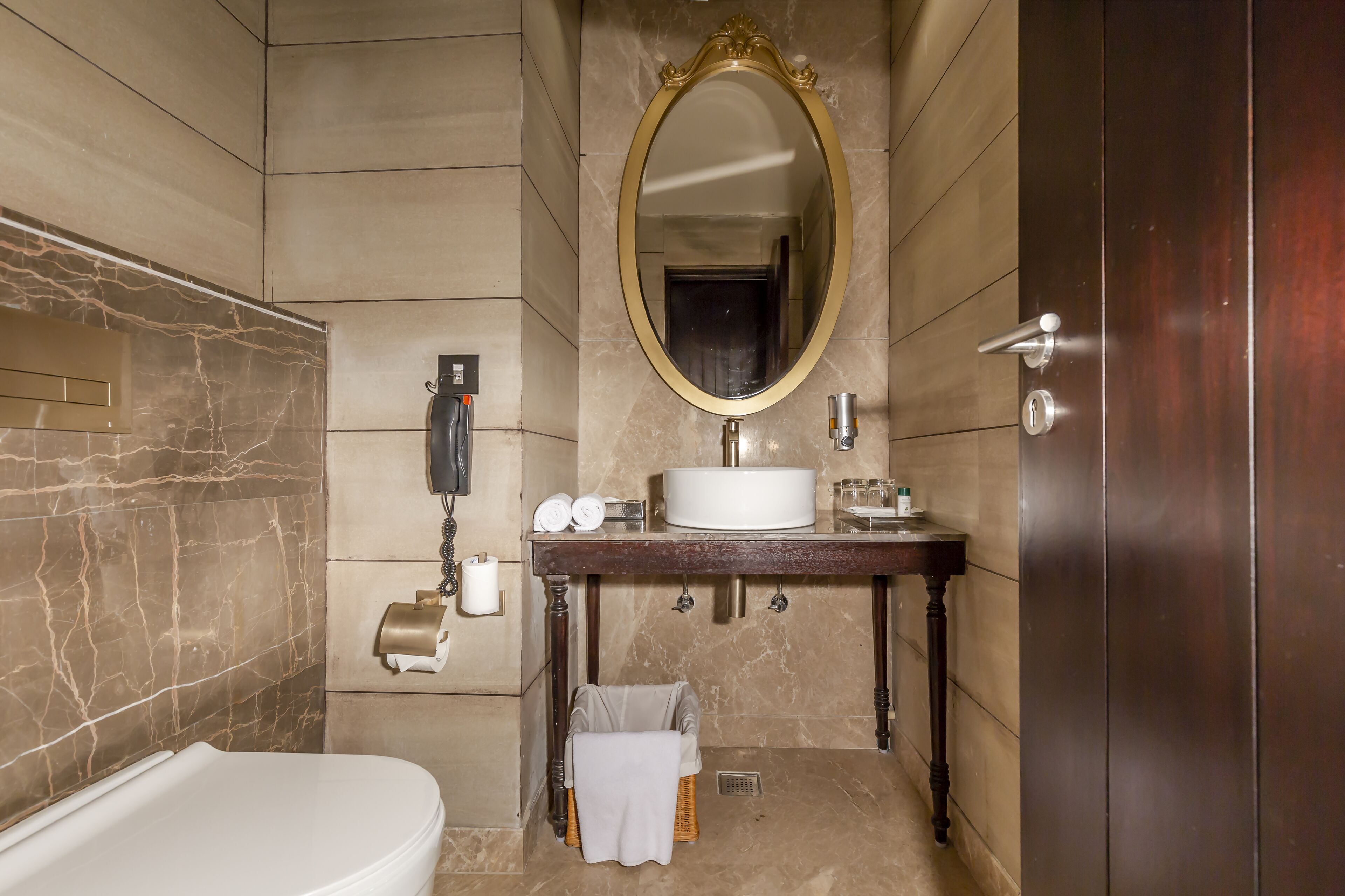 Un baño con pared de mármol, tocador de madera, un espejo grande, inodoro y toallero.