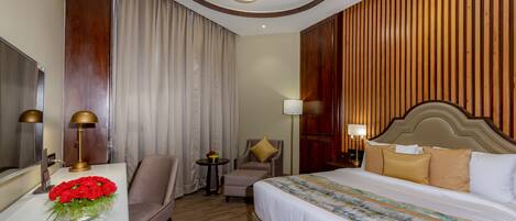 A hotel room with a large bed, a desk with a lamp, a chair, a small table with a vase of flowers, and a wooden wall paneling.