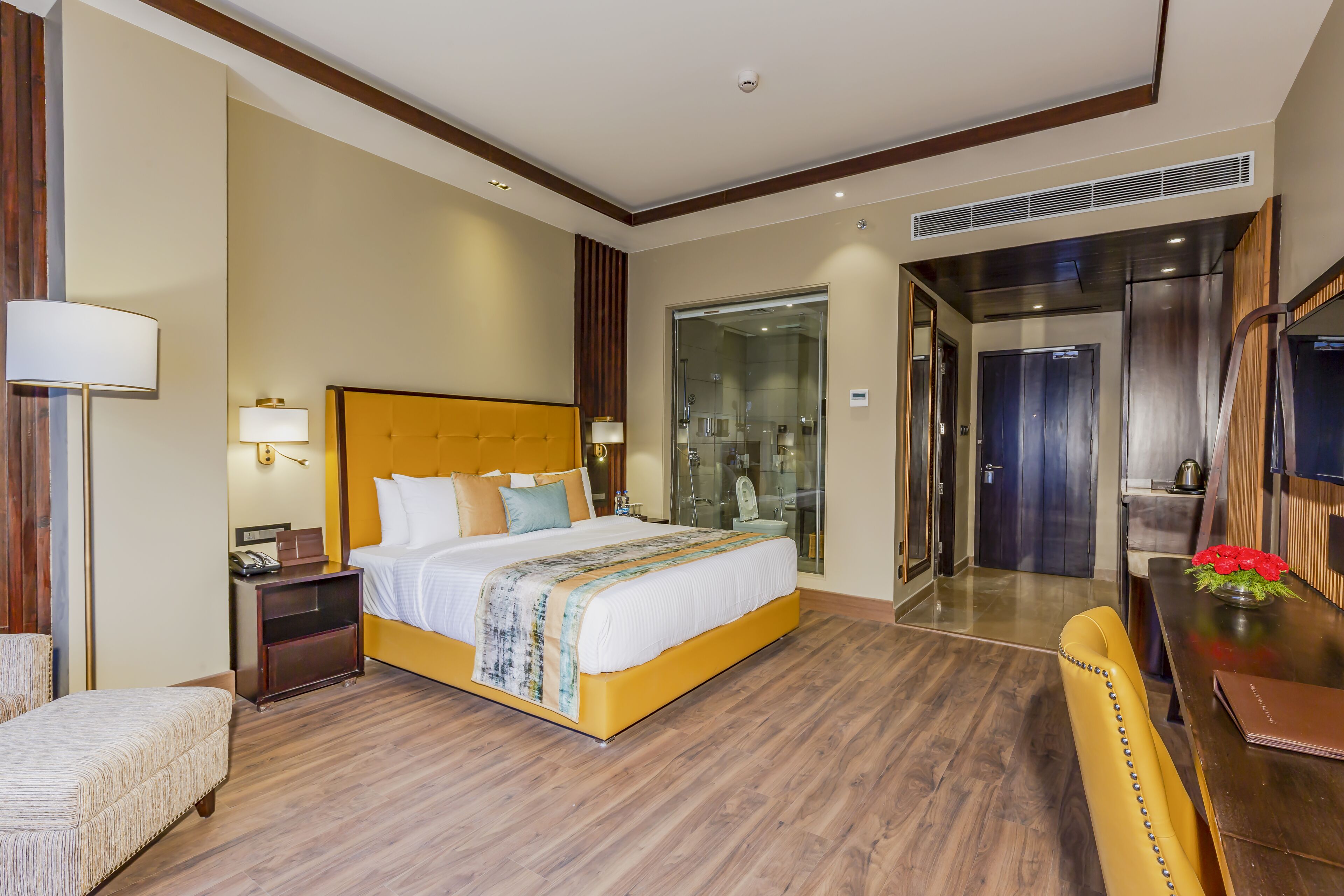 A hotel room with a large bed, bedside table, lamp, and a chair. There is a bathroom with a glass shower and a toilet visible through the door.