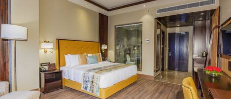 A hotel room with a large bed, bedside table, lamp, and a chair. There is a bathroom with a glass shower and a toilet visible through the door.