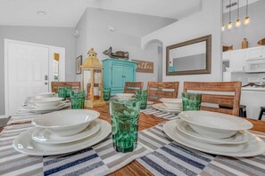Un comedor con una larga mesa dispuesta para seis personas, con individuales a rayas, platos blancos y vasos de vidrio verde. Se observa un aparador azul turquesa y una silla de madera al fondo.