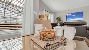 Una sala de estar moderna con una mesa de centro de vidrio, tazas de té blancas, un cuenco de vidrio con aperitivos, un sofá y un televisor que muestra una escena de playa.
