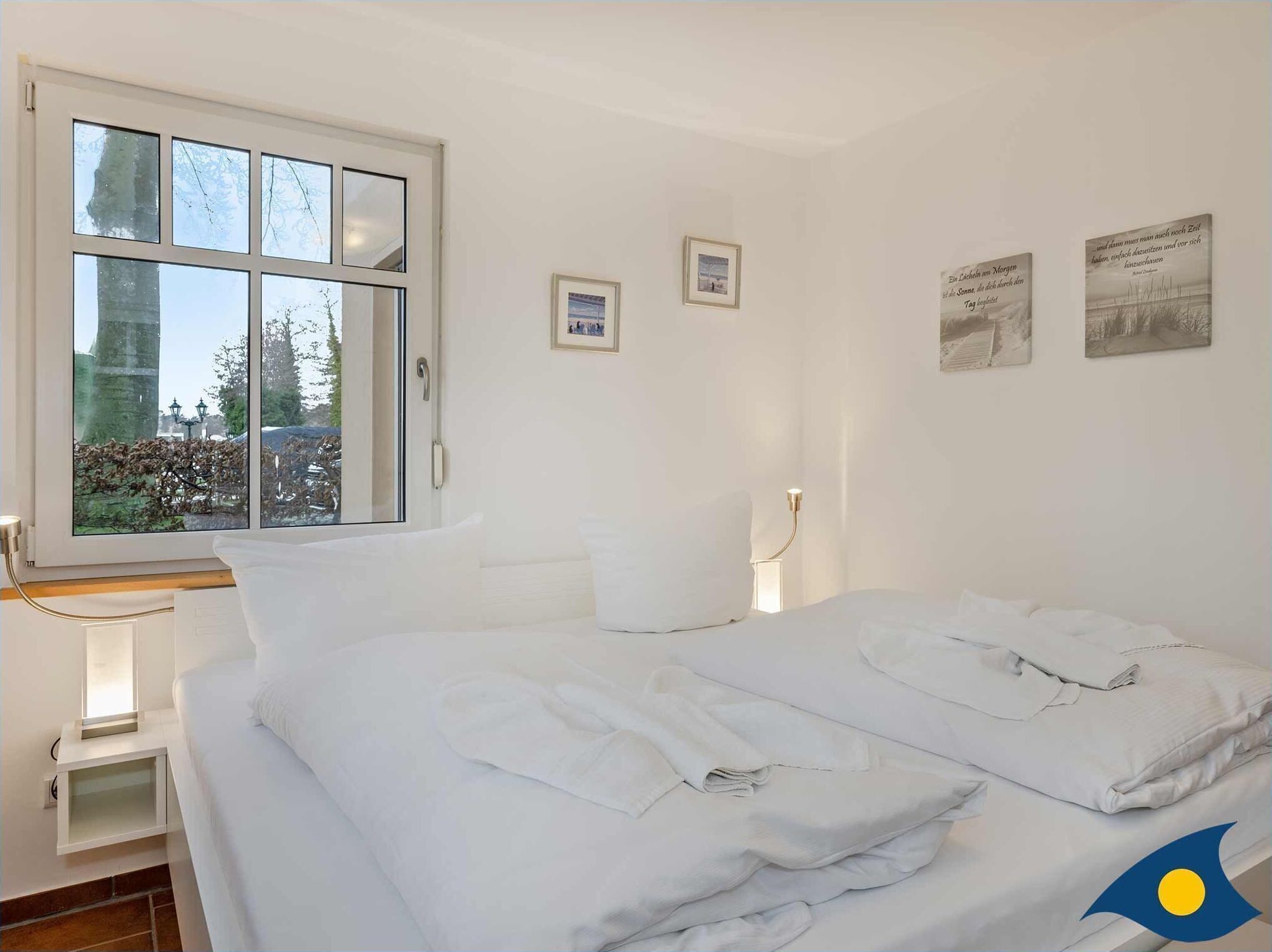 Une chambre d’hôtel avec un grand lit, une table de chevet, une fenêtre donnant sur des arbres et un immeuble, et deux tableaux encadrés accrochés au mur.