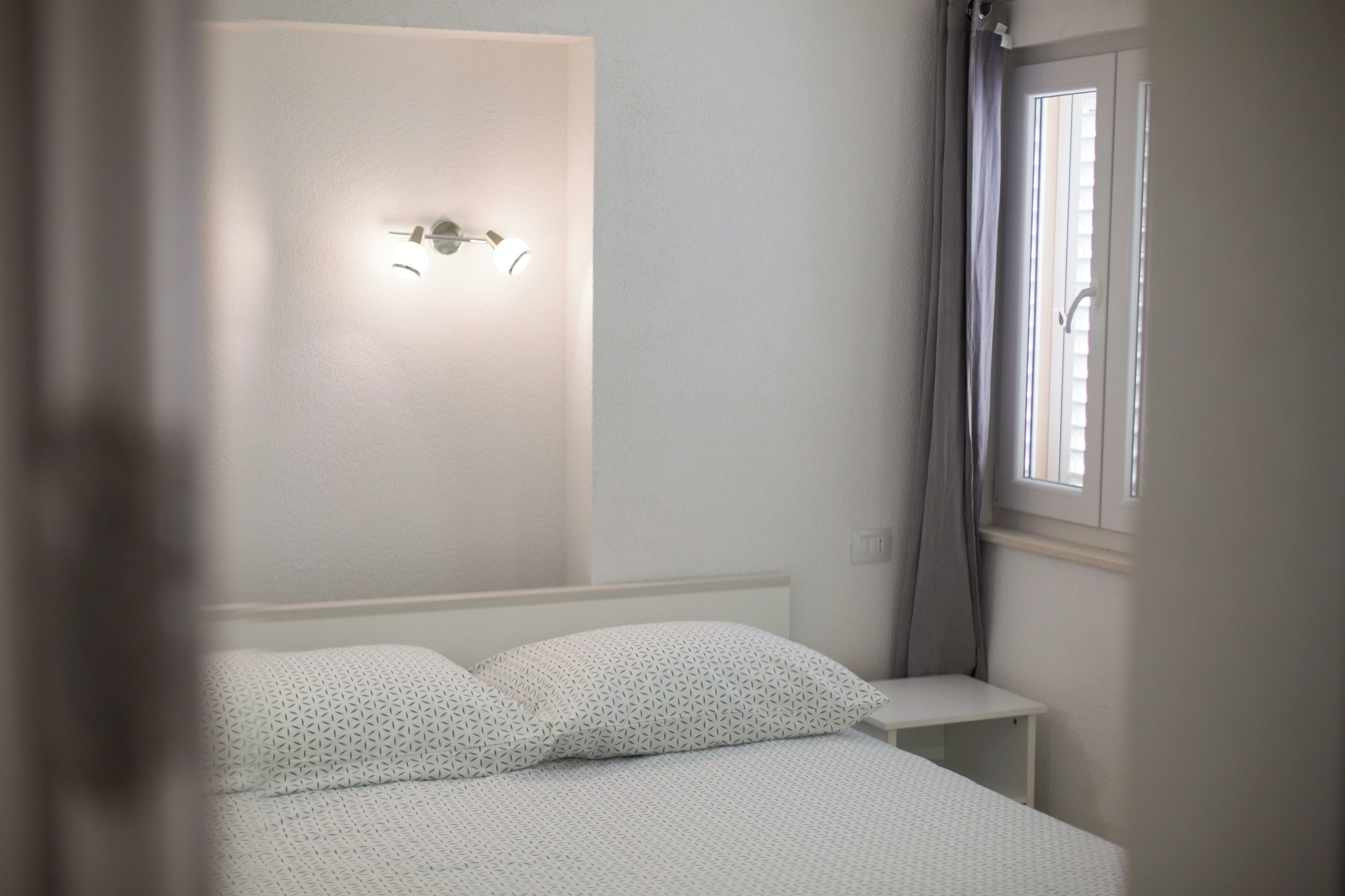 A bedroom with a bed, a nightstand, a wall-mounted light fixture, and a window with curtains.
