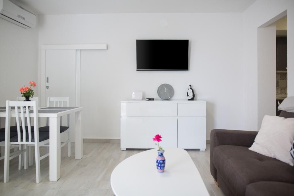 Un salon moderne avec un canapé blanc, une table à manger avec des chaises, un meuble blanc, un téléviseur à écran plat fixé au mur et un vase de fleurs posé sur une table basse.