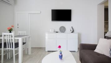 Un salon moderne avec un canapé blanc, une table à manger avec des chaises, un meuble blanc, un téléviseur à écran plat fixé au mur et un vase de fleurs posé sur une table basse.