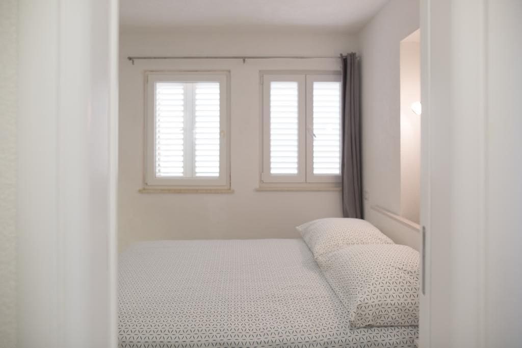 A bedroom with a single bed, two windows with blinds, and a light fixture above the bed.