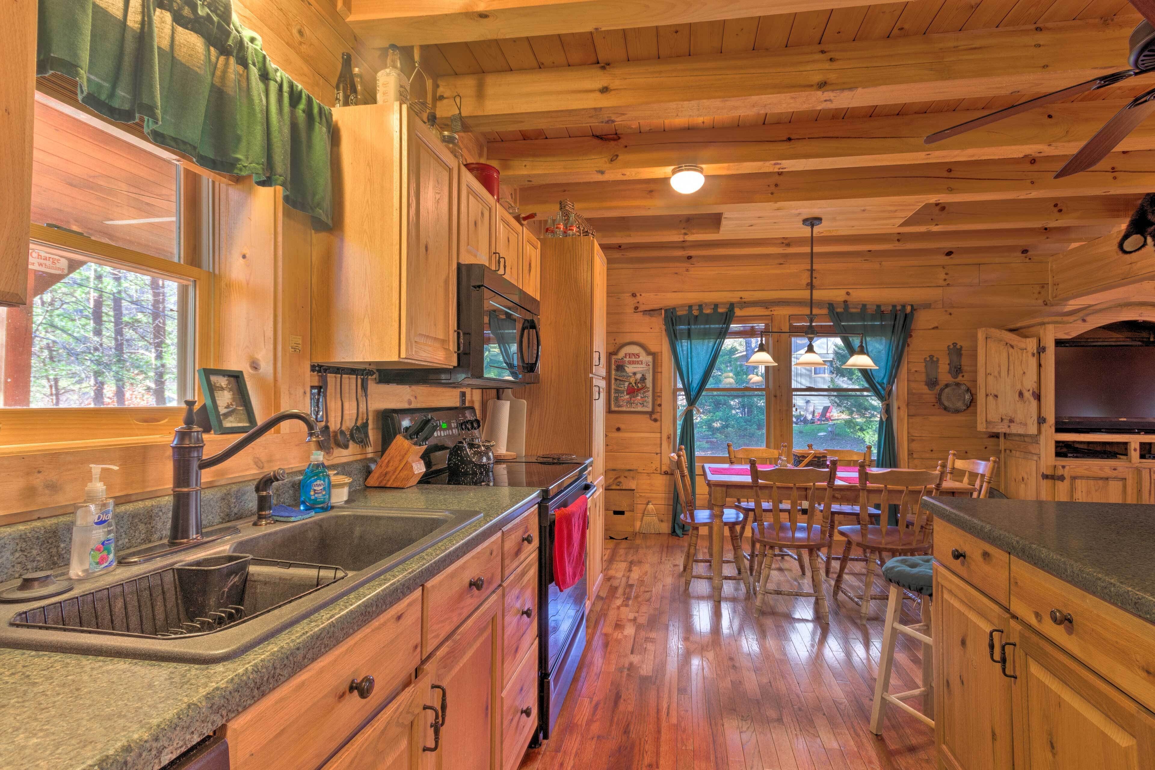 A wooden cabin interior with a kitchen, dining area, and a large window overlooking a forest.