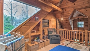 Ein gemütliches Schlafzimmer unter dem Dach mit geneigter Decke, Holzbalken, Deckenventilator, großem Fenster und Blick ins Freie.