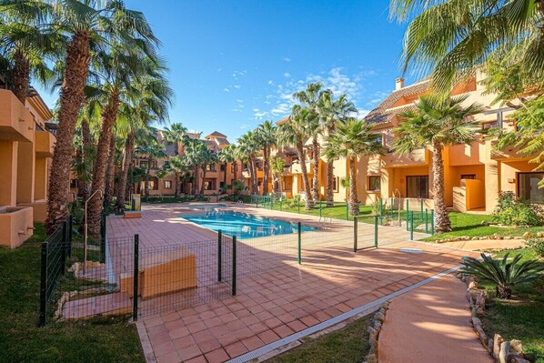 Un área de piscina con piscina cercada, rodeada de palmeras y edificios con fachadas amarillas y anaranjadas.