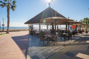 Un café al aire libre con mesas y sillas, un pabellón con techo de paja y vista al mar.