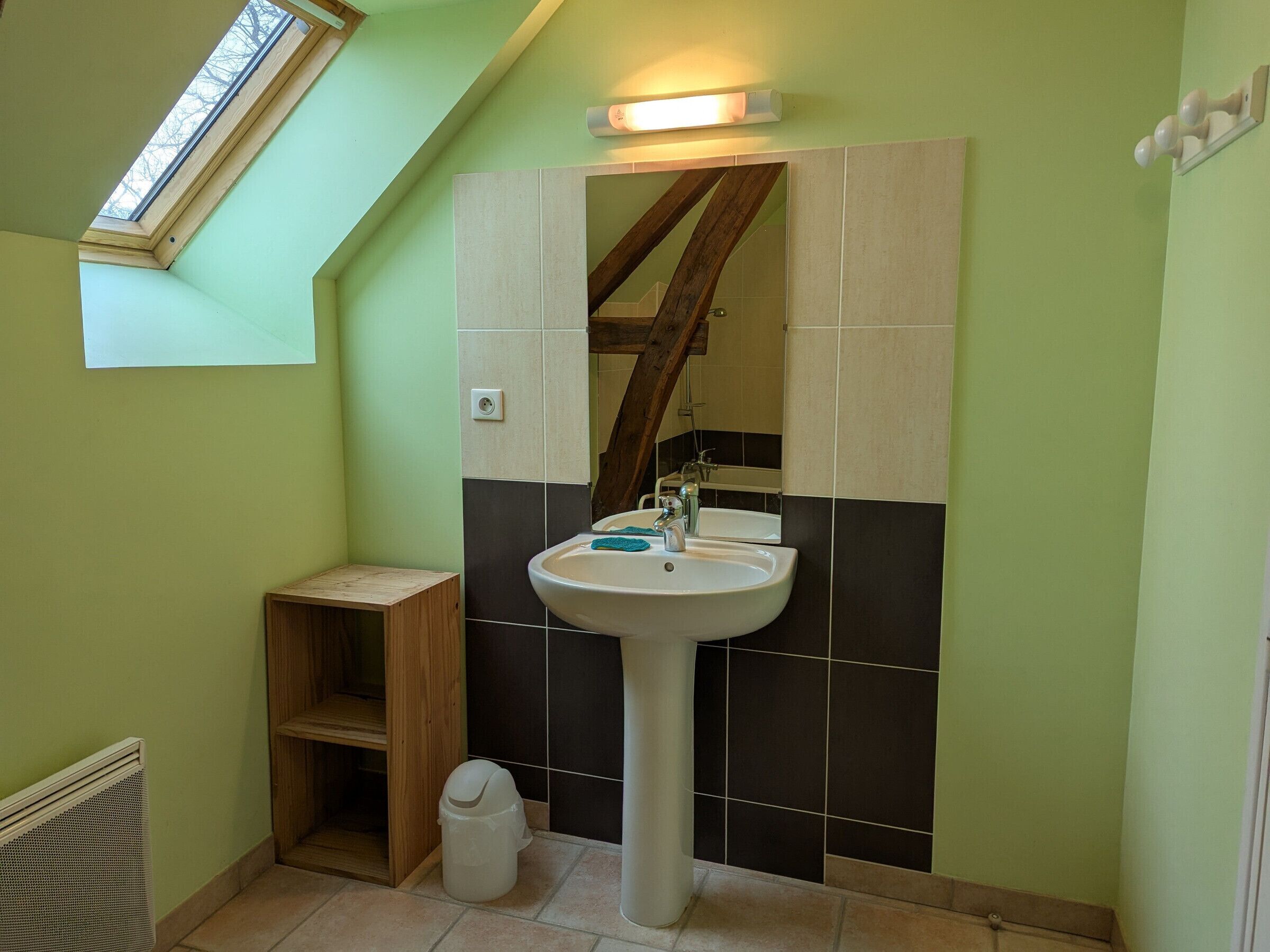 A bathroom with a skylight, a pedestal sink, a wooden shelf, and a trash bin.