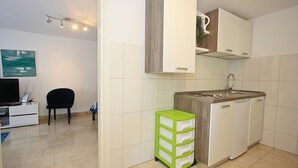 A compact kitchen with a dishwasher, a small sink, and a cabinet above the counter. There's a chair and a TV in the adjacent room. - Apartment Zorica (Supetar)