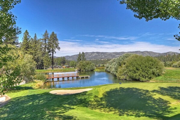 A golf course with a pond, wooden bridge, and mountains in the background.