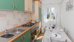 A compact kitchen with green cabinets, a gas stove, and a sink. A dining table with plates and glasses is set up near a window with white curtains. - Apartment Fiorella (Dubrovnik)