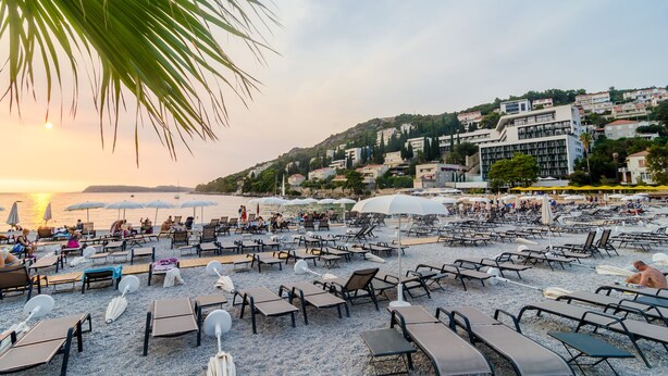 A beach with rows of sun loungers and umbrellas, a clear sky, and buildings in the background. - Apartment Fiorella (Dubrovnik)