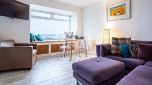 A modern living room with a sofa, a dining table with chairs, and a large window offering a view. - Headline (VRBO): Sunset View : Pet-Friendly 2‑Bed Apartment with Bay Views in Rhosneigr | Managed by (Rhosneigr)