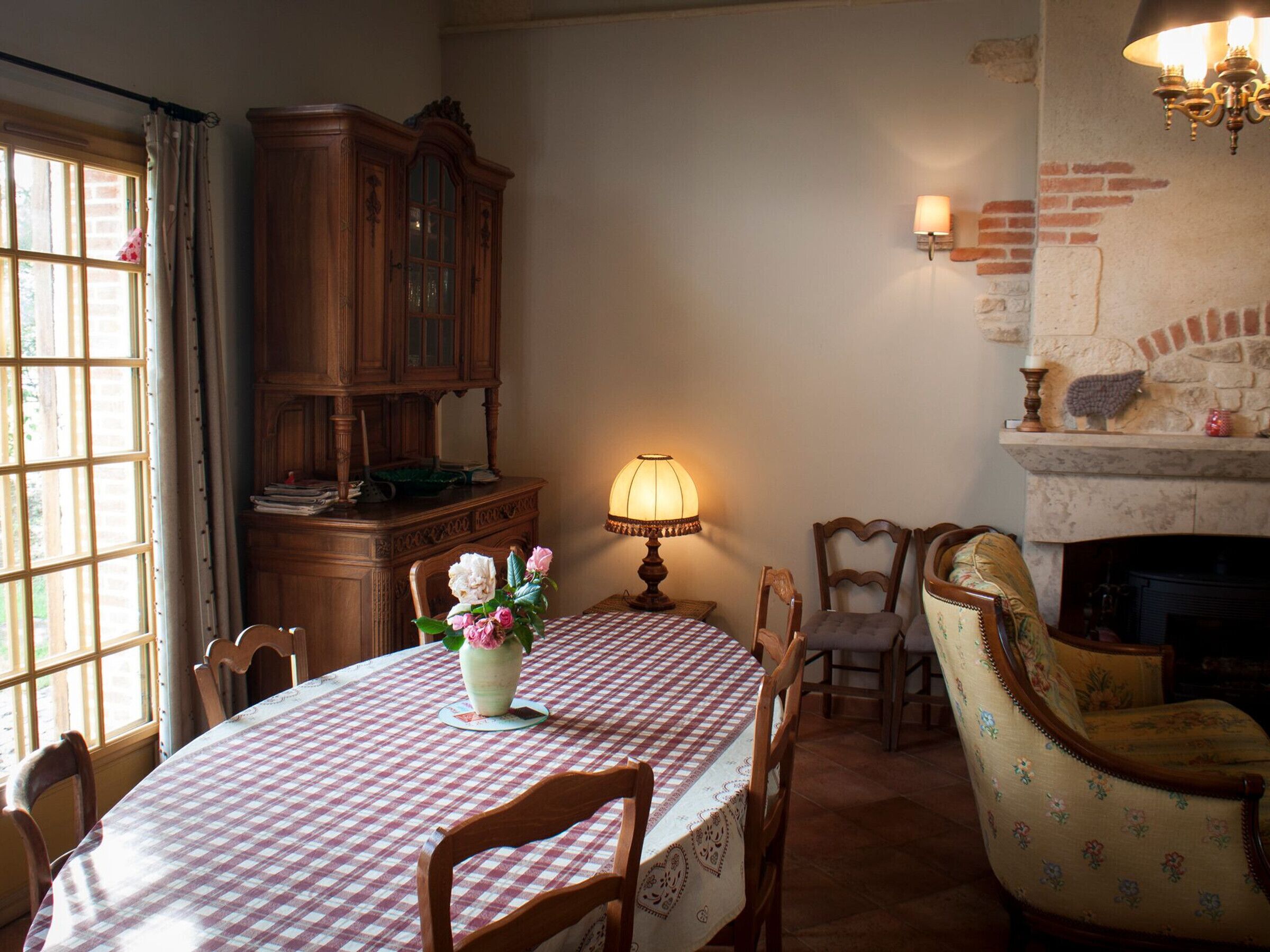 A dining room with a round table covered in a checkered tablecloth, surrounded by wooden chairs. A wooden cabinet and a lamp are also visible.