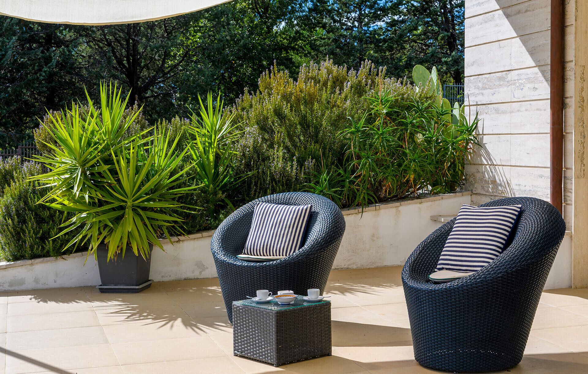 Dos sillones de mimbre con cojines a rayas y una pequeña mesa con tazas y un bol, ubicados en un patio empedrado con abundante vegetación y un muro de piedra.