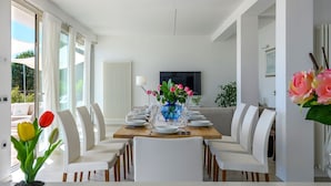 Ein Esszimmer mit einem langen Tisch, gedeckt für sechs Personen, weißen Stühlen, einer blauen Vase mit Blumen und einem Fernseher an der Wand.