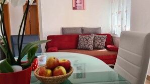 A living room with a glass table, a red sofa, a grey sofa, and a red armchair. There is a basket of apples on the table and a potted plant.