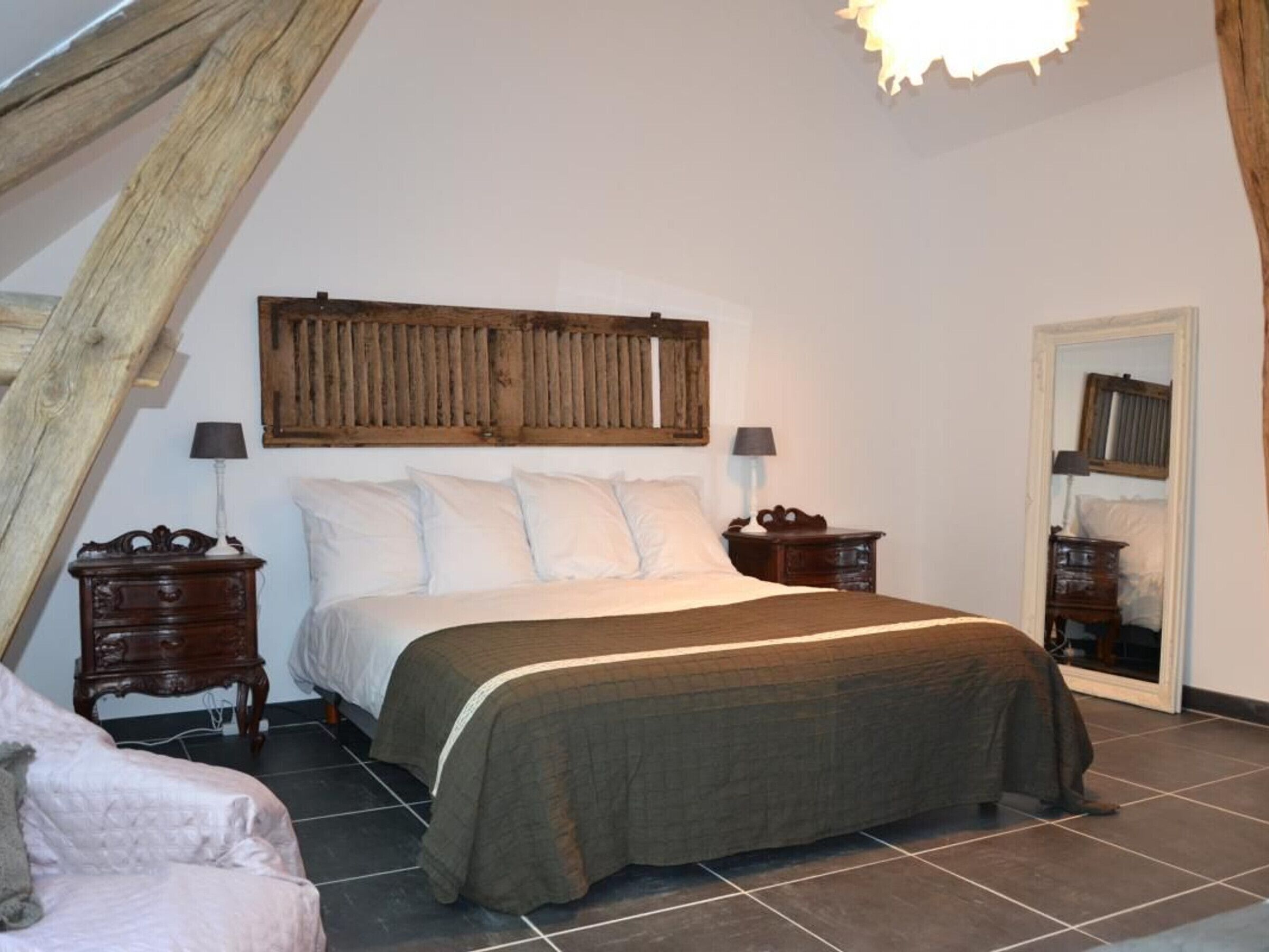Un dormitorio con techo inclinado, una cama con cabecera de madera, dos mesitas de noche con lámparas y un espejo en la pared.