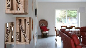 Un intérieur moderne avec des boîtes de rangement en bois fixées au mur, portant les inscriptions « HAUT » et « BAS », un canapé en cuir rouge et un coin repas avec des chaises et une table.