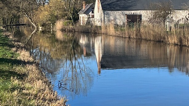 Une vue sur la rivière, avec un bâtiment en pierre, une cheminée et son reflet dans l’eau.