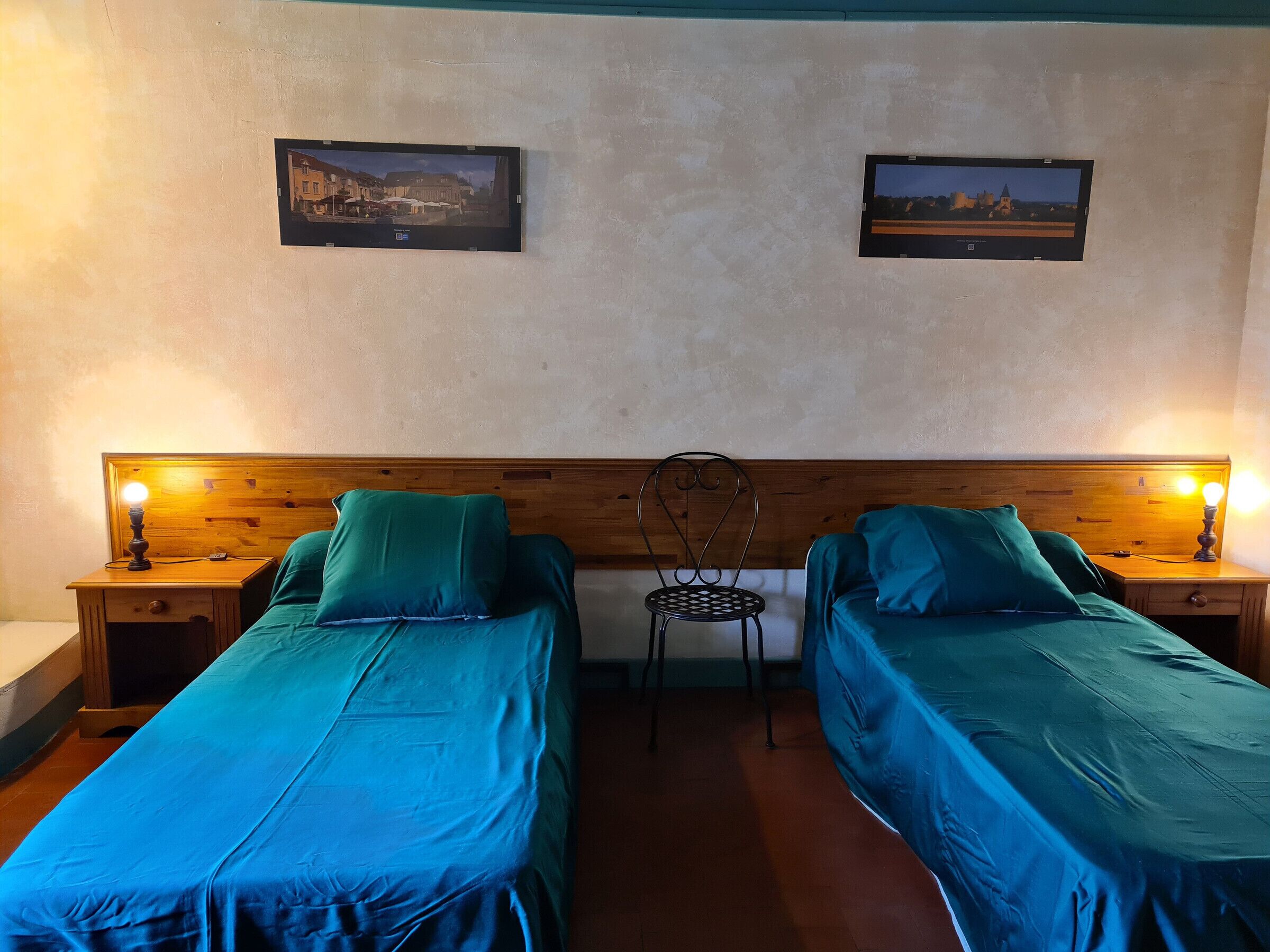 A room with two beds, each with a wooden headboard and a nightstand with a lamp. There are two framed pictures on the wall.