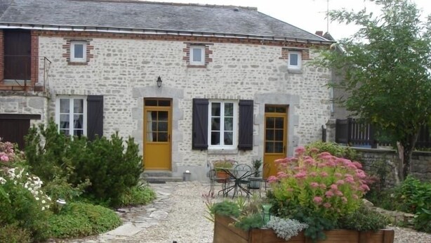 Un bâtiment en pierre avec un jardin à l’avant, comprenant une charrette en bois remplie de plantes.