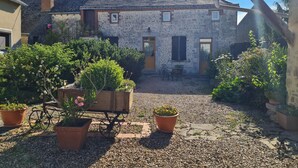 Un bâtiment en pierre avec une cour recouverte de gravier, entouré de plantes en pot et d’un chariot en bois.