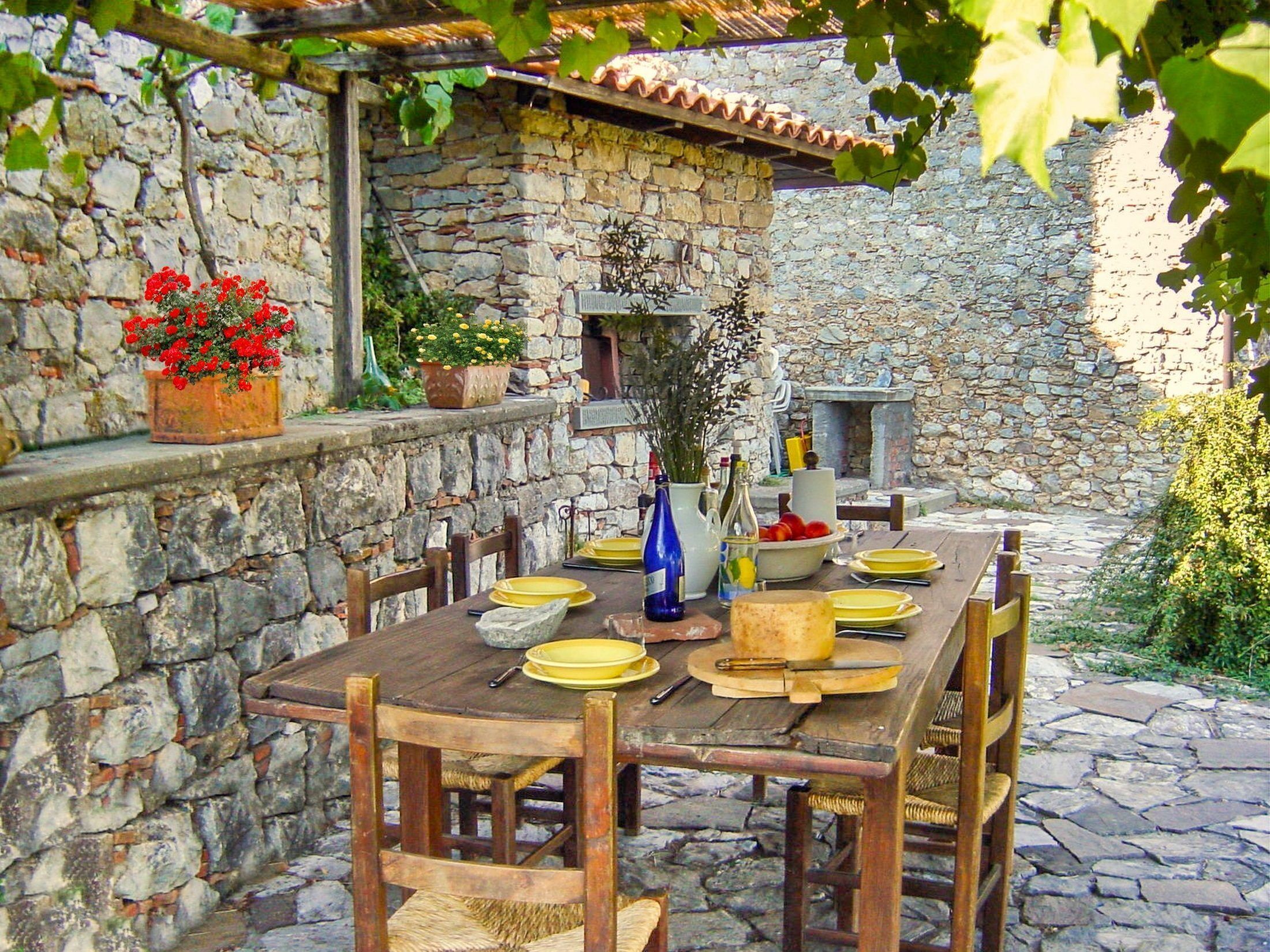 Un bâtiment rustique en pierre, avec une table en bois dressée pour le repas, entouré de murs en pierre et de plantes en pot.