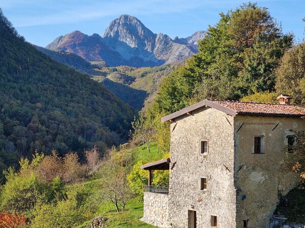 Um edifício rústico de pedra com telhado inclinado, cercado por vegetação exuberante e montanhas ao fundo.