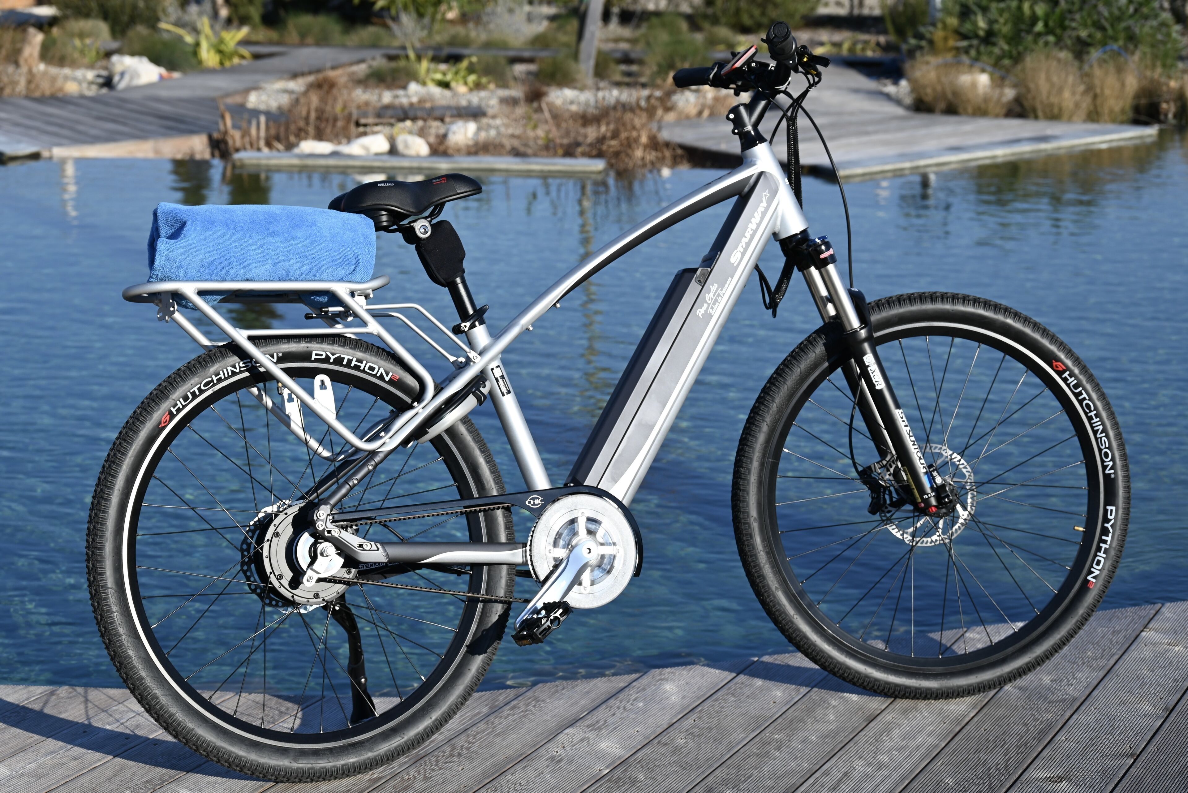 Una bicicleta elèctrica immobilitzada amb una tovallola blava al seient, un cisteller i un suport per ampolla d'aigua, aparcada sobre un moll de fusta al costat d'una massa d'aigua.