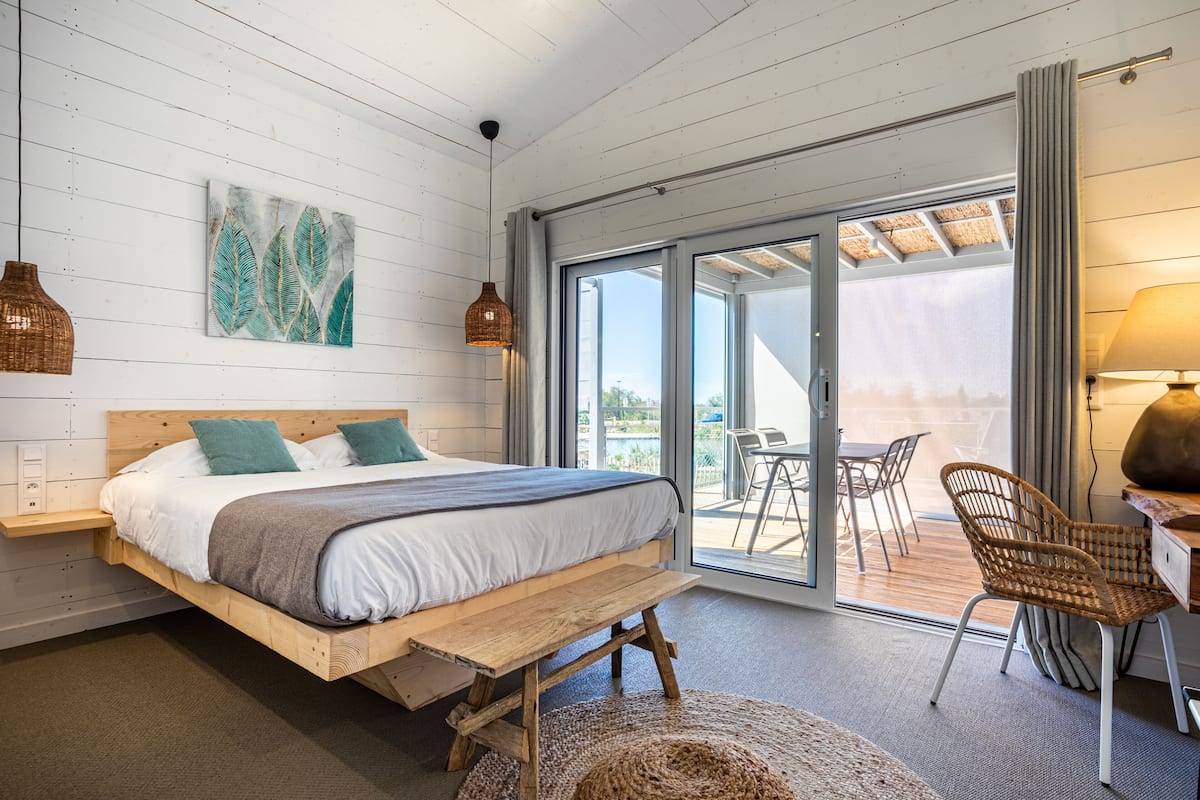 Une chambre à coucher avec un lit en bois, un banc, une chaise en osier et une vue sur l’extérieur grâce à de grandes portes coulissantes.
