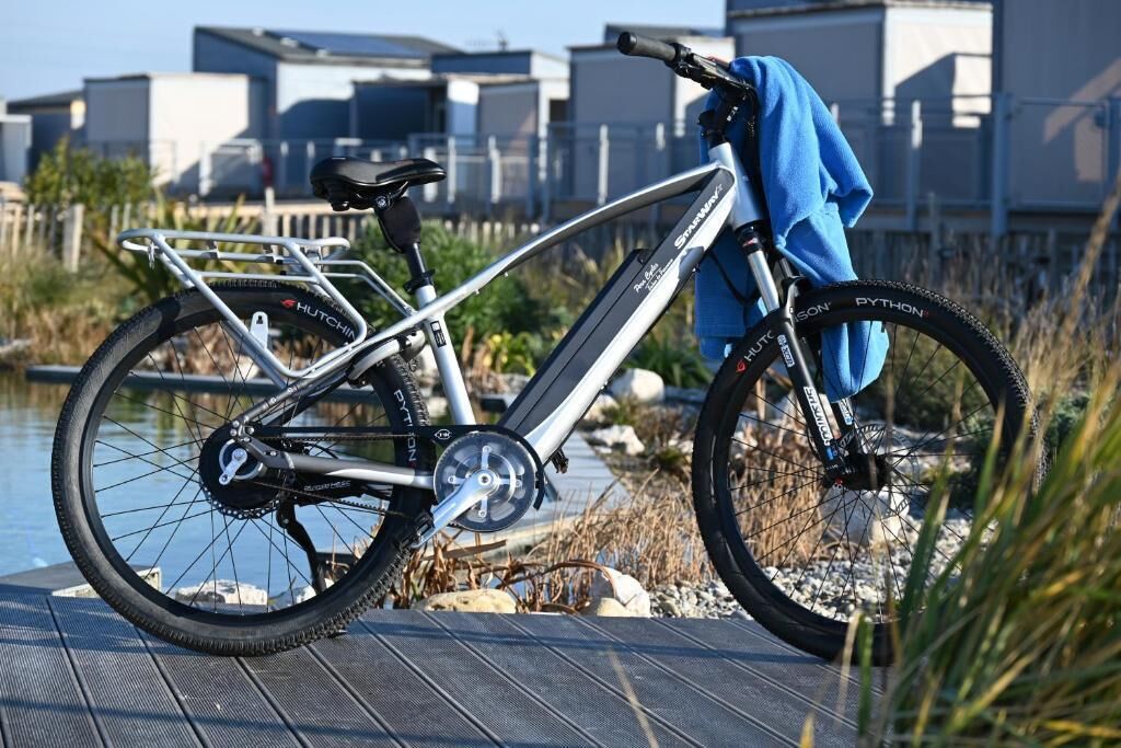 Ein stehendes Fahrrad mit einer Wasserflasche und einem Handtuch auf dem Sitz, abgestellt auf einer Holzterrasse in der Nähe eines Gewässers.